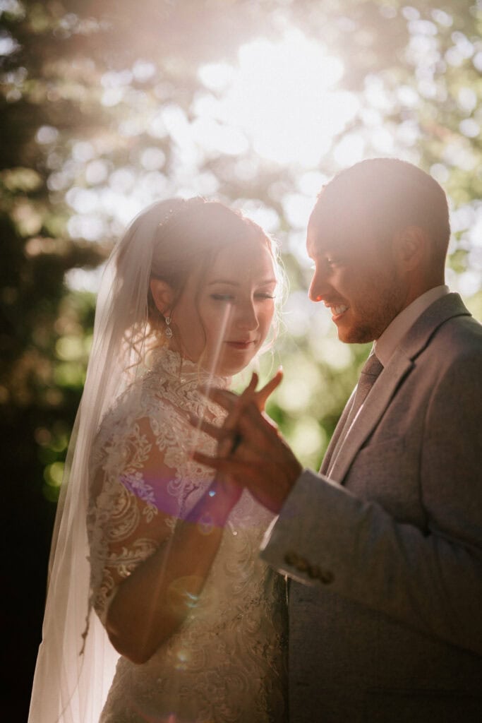 A bride in a lace dress and veil stands with a groom in a light suit at The Plough at Leigh, holding hands in a sunlit outdoor setting. The soft sunlight filters through the trees, creating a warm, romantic wedding atmosphere. Image by Pearce Wedding Photography.