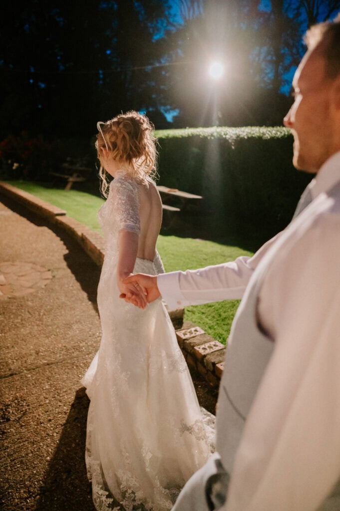 A bride and groom are holding hands, walking outside The Plough at Leigh in the evening. The bride dazzles in a white, open-back dress; the groom is sharp in a light-colored suit. Their wedding setting glows with soft lights, with a charming garden as the backdrop. Image by Pearce Wedding Photography.