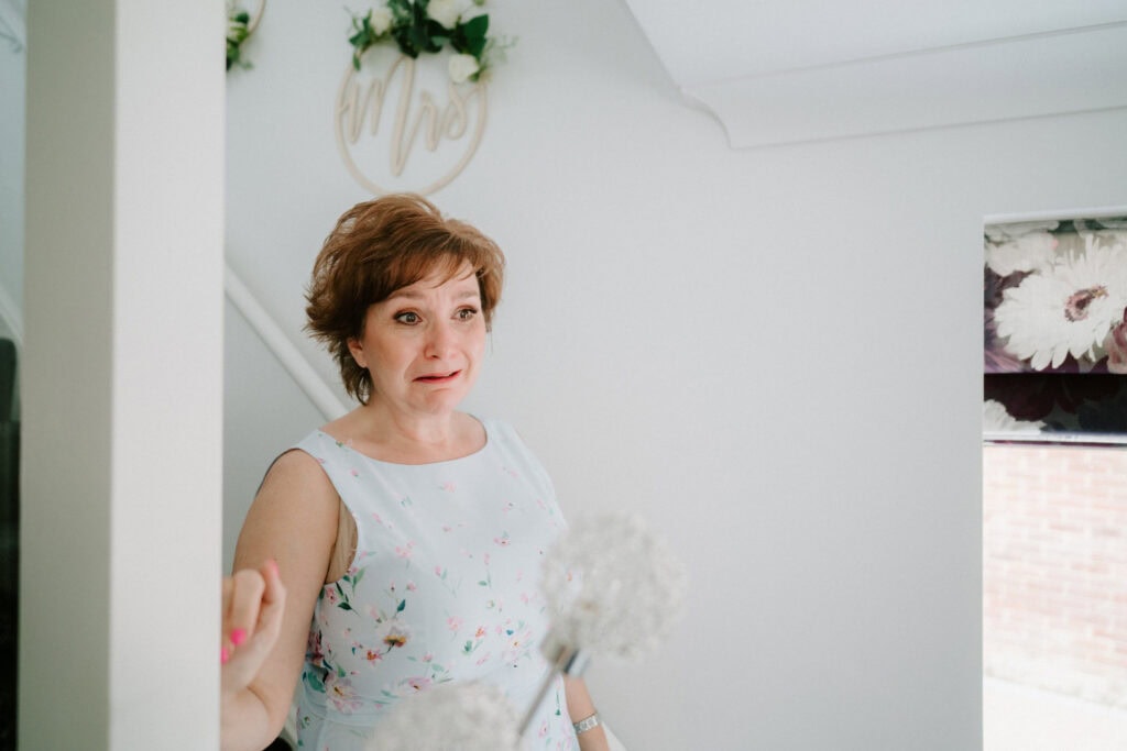 A woman in a floral dress stands near a staircase at The Plough at Leigh, looking emotional. Above her, a beautiful floral decoration sets the scene, capturing the essence of an unforgettable wedding day. Image by Pearce Wedding Photography.