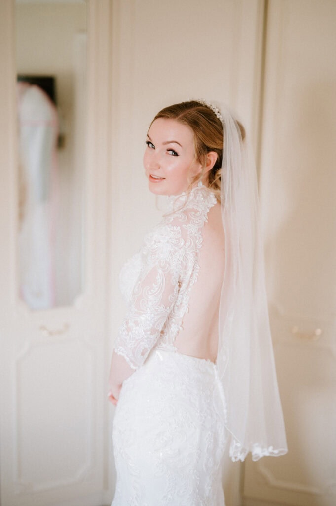A bride in a white lace wedding dress with an open back and long sleeves smiles over her shoulder. She is wearing a veil and standing in front of a cream-colored wardrobe, capturing a moment of pure joy before joining her guests at The Plough at Leigh for the wedding celebration. Image by Pearce Wedding Photography.