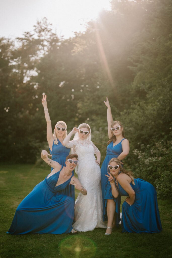 A bride in a white dress poses outdoors at The Plough at Leigh with four bridesmaids in blue dresses. They all wear heart-shaped sunglasses and make playful poses, with greenery and sunlight framing this delightful wedding moment. Image by Pearce Wedding Photography.