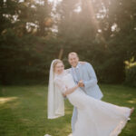 A bride in a white wedding dress and veil is joyfully held by a groom in a light gray suit on the sunlit lawn of The Plough at Leigh. Surrounded by trees, with sunlight filtering through, they are enveloped in a warm and romantic wedding atmosphere. Image by Pearce Wedding Photography.
