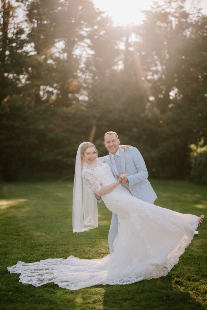 A bride in a white wedding dress and veil is joyfully held by a groom in a light gray suit on the sunlit lawn of The Plough at Leigh. Surrounded by trees, with sunlight filtering through, they are enveloped in a warm and romantic wedding atmosphere. Image by Pearce Wedding Photography.