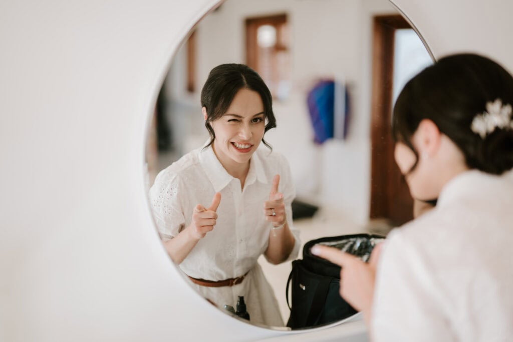 In a softly focused room at Pretty Prep Studios Ashford, a woman with dark hair styled in a low bun decorated with a hairpin smiles and winks at her reflection in a round mirror. She's wearing a white button-up shirt and playfully pointing her fingers like pretend guns. Image by Pearce Wedding Photography.
