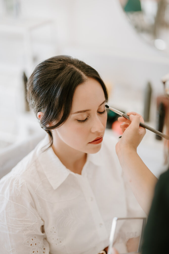 A woman with dark hair is having her makeup meticulously applied by another person. Dressed in a white blouse, she sits with her eyes closed in what appears to be a makeup studio filled with various beauty products. The scene evokes the careful preparations before a wedding at "The Secret Garden" in Kent. Image by Pearce Wedding Photography.