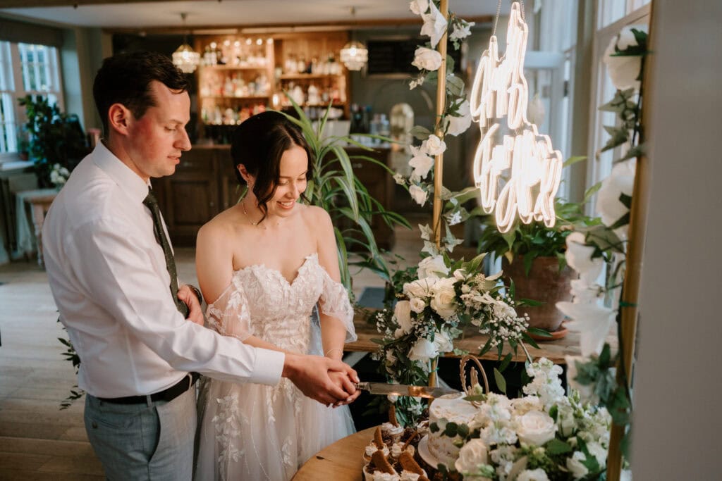 A couple, dressed in wedding attire, smiles as they cut their wedding cake together at The Secret Garden in Kent. The bride wears a white off-shoulder gown with lace details, and the groom is in a white shirt and tie. The cake table is decorated with flowers and greenery, and a neon sign glows in the background. Image by Pearce Wedding Photography.