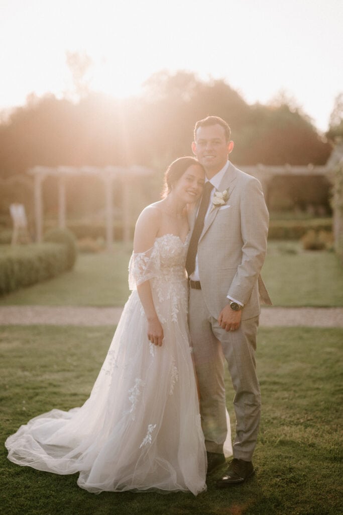 A couple stands closely together in a garden at sunset in Kent. The woman is wearing a flowing white wedding dress with lace details and off-the-shoulder sleeves, while the man is dressed in a light grey suit with a boutonniere. They are smiling softly, embracing, and savoring their magical wedding moment. Image by Pearce Wedding Photography.