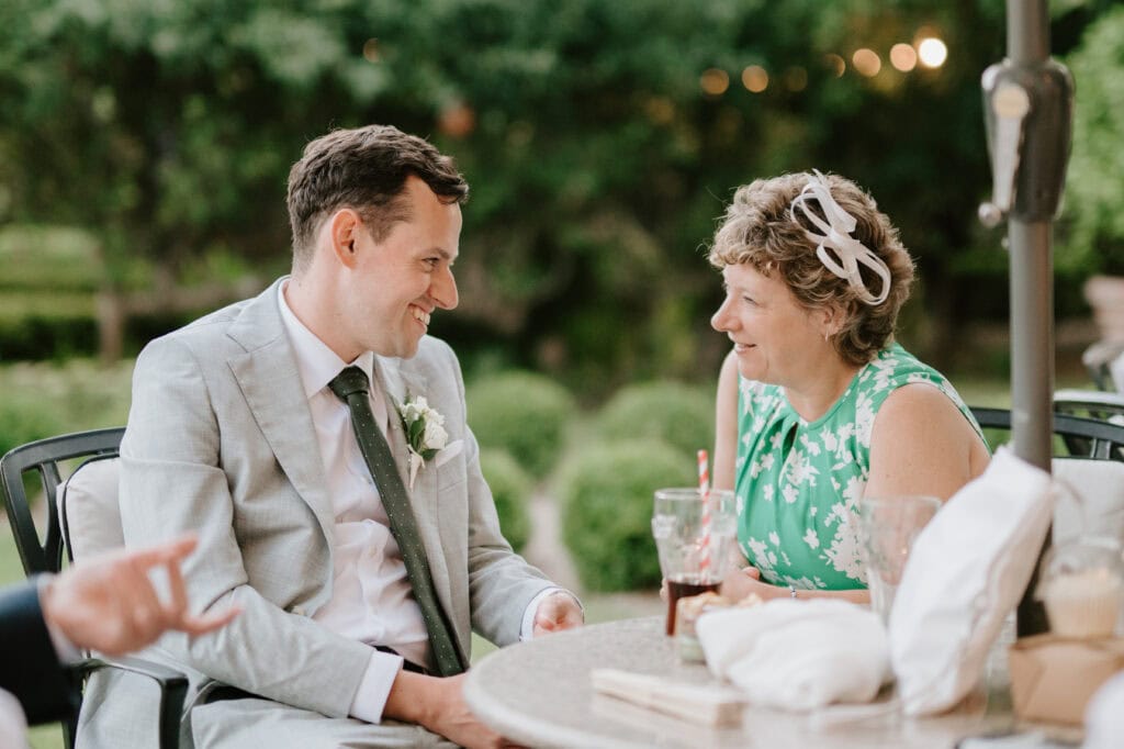 A man in a light gray suit with a white flower boutonniere is smiling while sitting at a table with a woman in a green floral dress. They are outdoors, resembling Kent's lush landscapes from "The Secret Garden," and there are drinks and tableware before them, suggesting it's either a lovely luncheon or possibly even during a wedding celebration. Image by Pearce Wedding Photography.