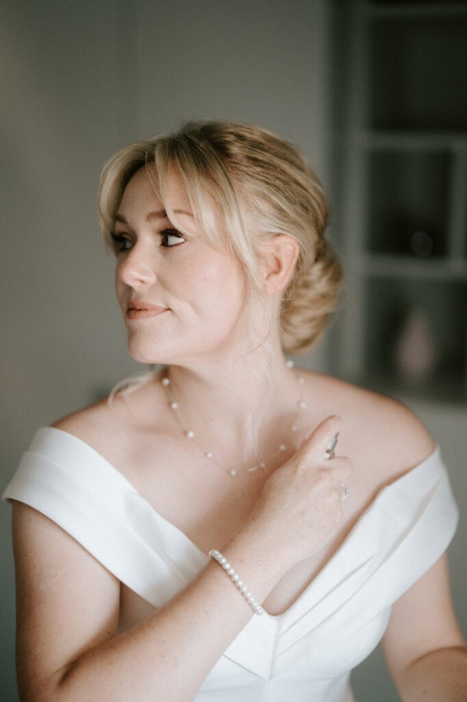 A woman with blonde hair, styled in an updo, stands sideways in a well-lit room at her Whitstable wedding. She is wearing an off-shoulder white dress, a pearl necklace, and a matching bracelet. With a subtle smile, she raises her hand to her chest. The background is softly blurred. Image by Pearce Wedding Photography.