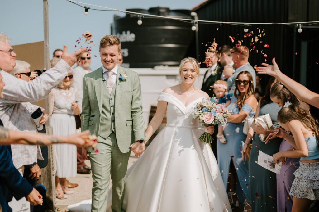 A newlywed couple, the groom in a light green suit and the bride in a white gown, walk hand in hand as guests throw flower petals. They are both smiling, and the bride holds a colorful bouquet. The outdoor wedding celebration at East Quay in Whitstable is decorated with string lights. Image by Pearce Wedding Photography.