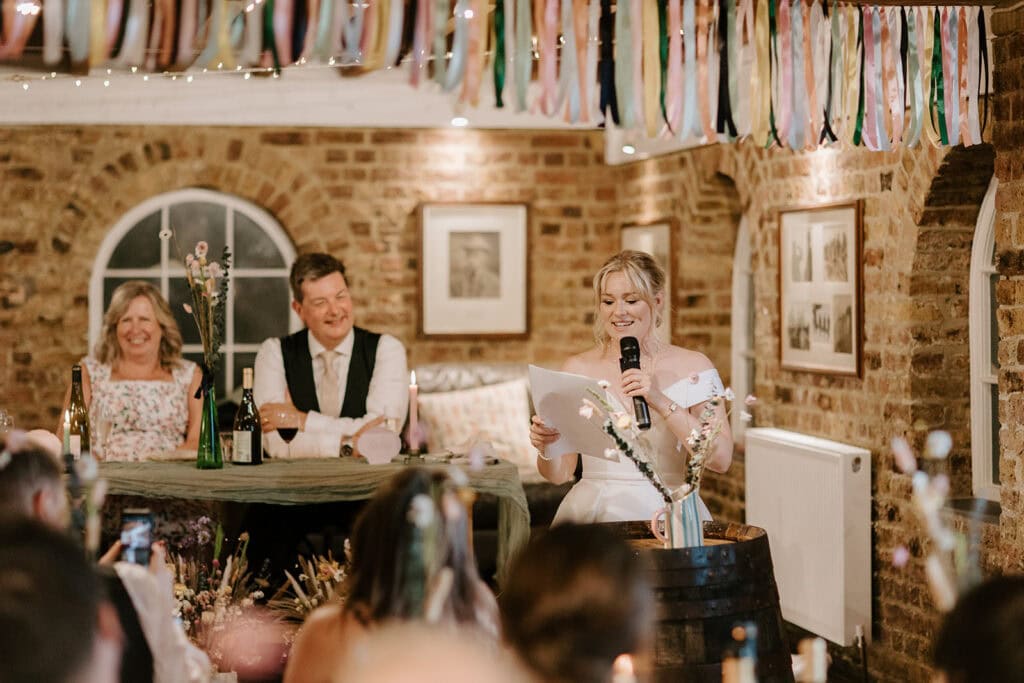 At the East Quay venue, a woman in a white dress delivers a heartfelt wedding speech indoors, microphone and notes in hand. A man and woman sit smiling at a table adorned with bottles and flowers. Colorful ribbons hang above, set against brick walls and framed pictures. Image by Pearce Wedding Photography.