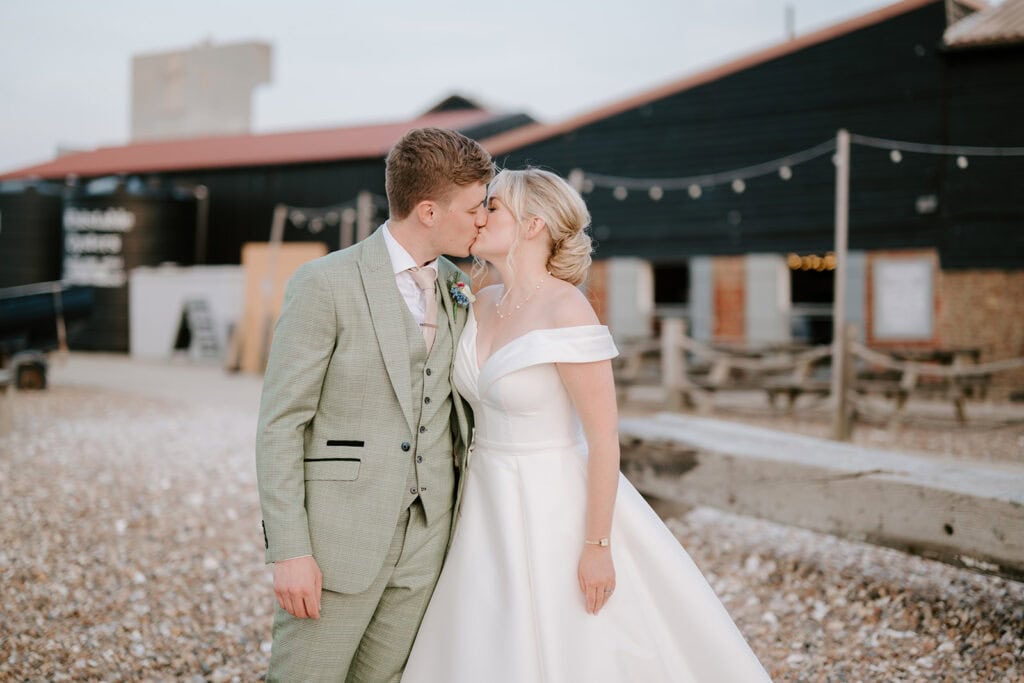 A bride in an off-shoulder white gown and a groom in a light gray suit share a kiss outside East Quay Venue. They stand on a rocky path, with the rustic charm of the barn-like building and twinkling string lights creating the perfect wedding backdrop. Image by Pearce Wedding Photography.