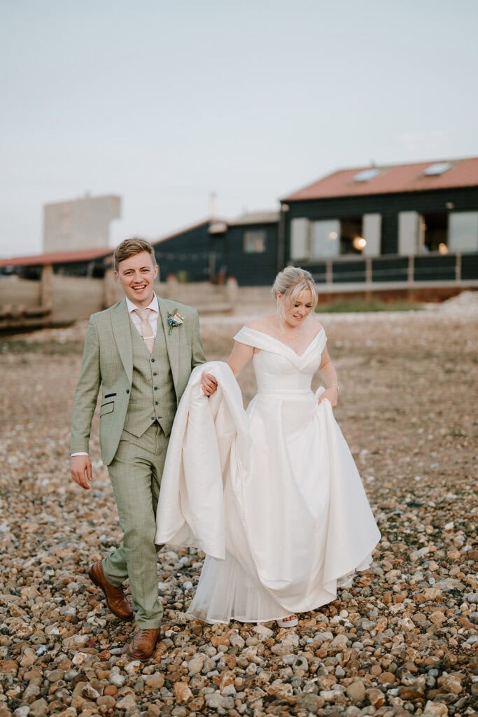 A bride and groom stroll along a rocky beach near the East Quay venue. The groom, in a light green suit, gently holds part of the bride's white dress. She smiles warmly in her off-the-shoulder gown, with rustic buildings set against a clear sky as their wedding backdrop. Image by Pearce Wedding Photography.