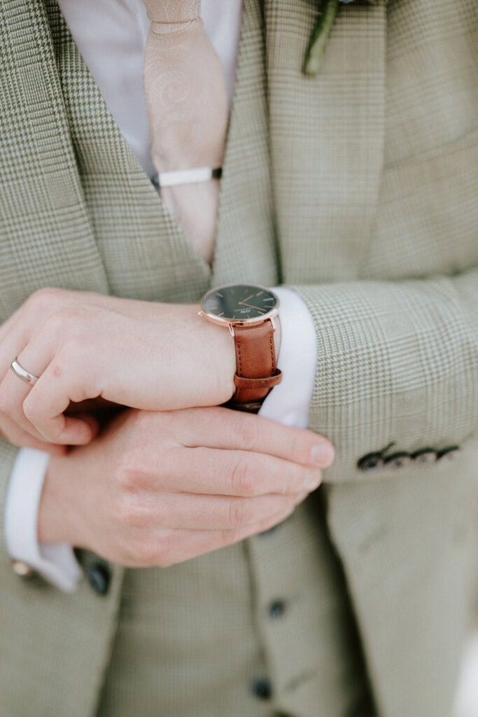 A person in a plaid suit adjusts a brown leather wristwatch, perhaps preparing for an event at the East Quay Venue. The focus is on the hands, suit, and tie, with a blurred background hinting at the excitement of a wedding day. Image by Pearce Wedding Photography.