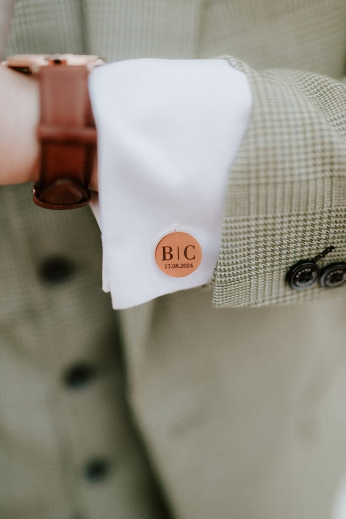 Close-up of a person wearing a light green checkered suit with a white shirt, perfect for a wedding at East Quay Venue. The cufflink features engraved initials "BIC" and the date "17.08.2024" in black lettering, while the wrist is adorned with a brown strap watch. Image by Pearce Wedding Photography.