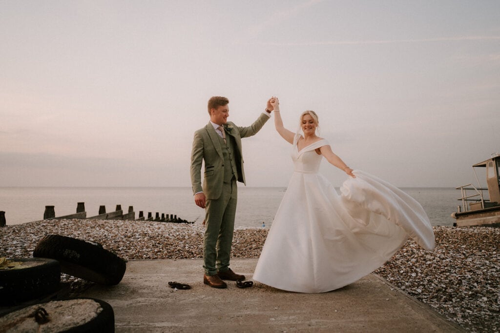 As the sun dips below the horizon, a couple dances on a pebble beach, their silhouettes framed by the shimmering sea. The woman, in a flowing white gown, and her partner in a light green suit share a quiet moment reminiscent of an intimate wedding at East Quay Venue amidst wooden beams and tires. Image by Pearce Wedding Photography.