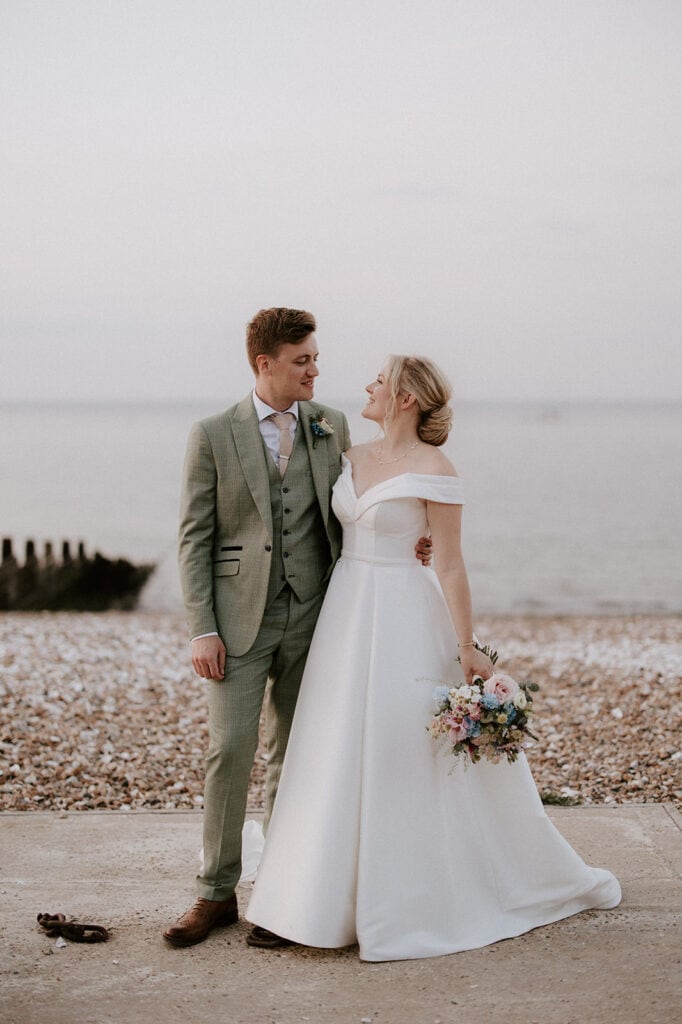 A bride and groom stand on a pebbled beach at East Quay Venue, gazing at each other and smiling. The groom wears a light grey suit, and the bride wears an off-shoulder white gown, holding a bouquet of flowers. The calm sea in the background completes the perfect wedding scene under a light sky. Image by Pearce Wedding Photography.