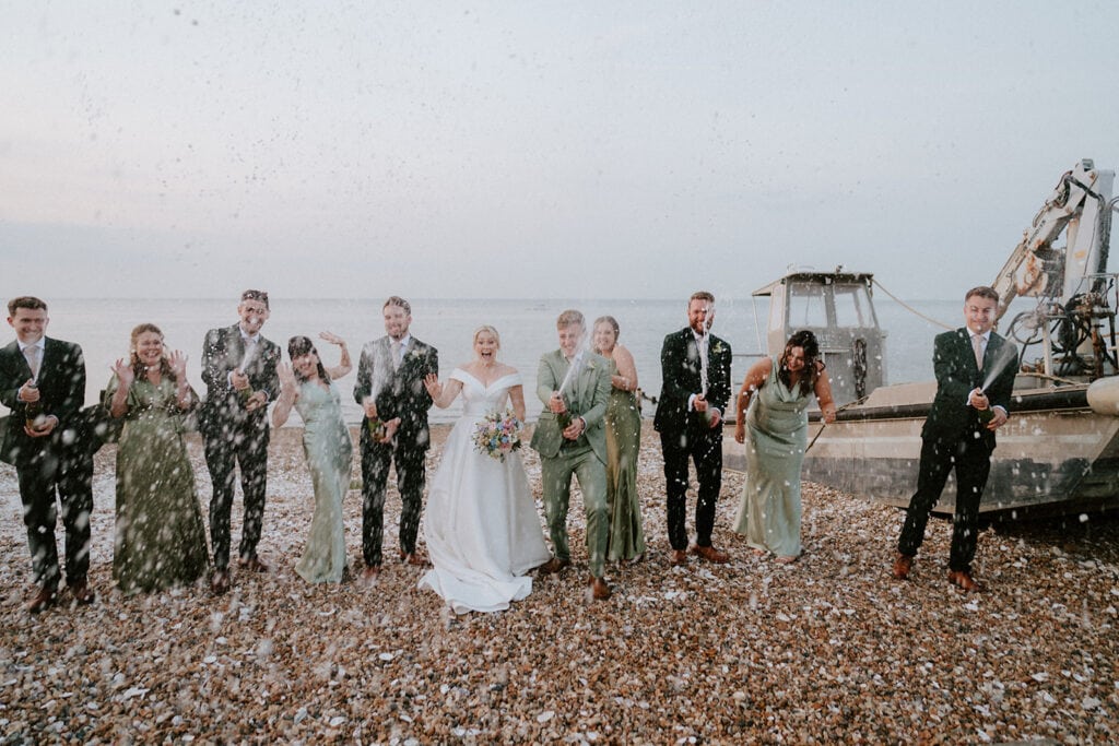 A wedding party stands on a pebbled beach, joyfully spraying champagne at the East Quay Venue. The bride, in her white dress, and the groom, sharply suited, are surrounded by bridesmaids and groomsmen in matching attire, with the sea and a boat creating a picturesque backdrop. Image by Pearce Wedding Photography.