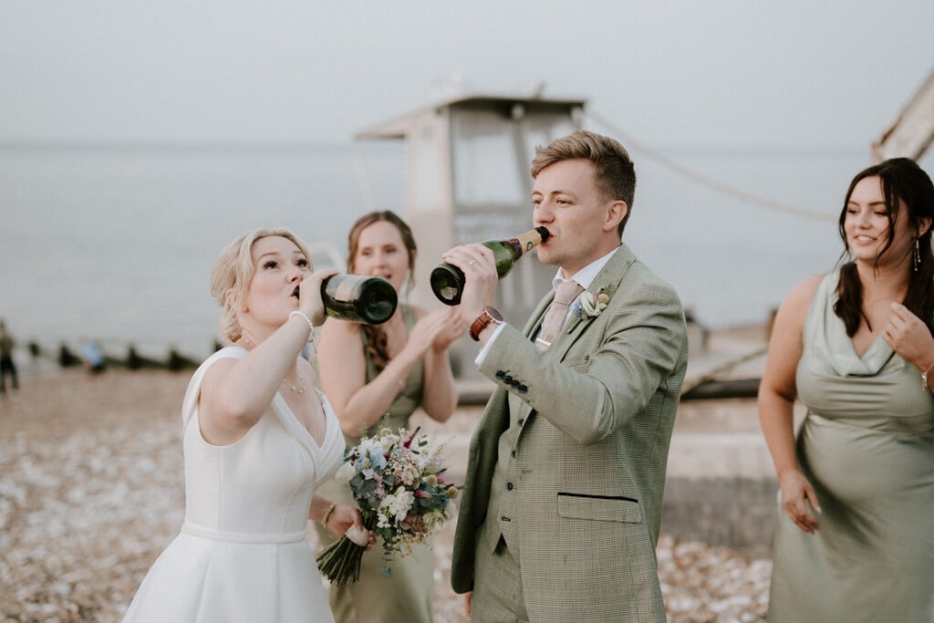 A bride and groom drink from bottles on a pebble beach, celebrating their wedding with three bridesmaids. The groom, in a green suit, and the bride, holding her bouquet, enjoy the stunning backdrop of a boat on the sea near the East Quay Venue. Image by Pearce Wedding Photography.