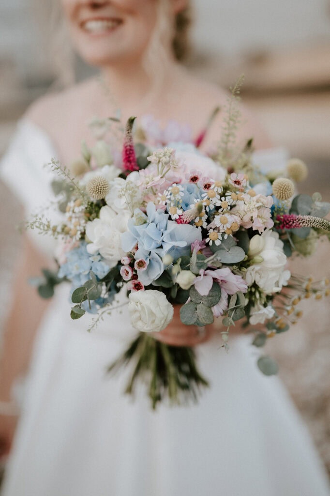 At the picturesque East Quay Venue, a bride in a white dress holds a vibrant wedding bouquet featuring blue hydrangeas, white roses, pink flowers, and greenery. The focus is on the bouquet, with her smiling face slightly blurred in the background. Image by Pearce Wedding Photography.