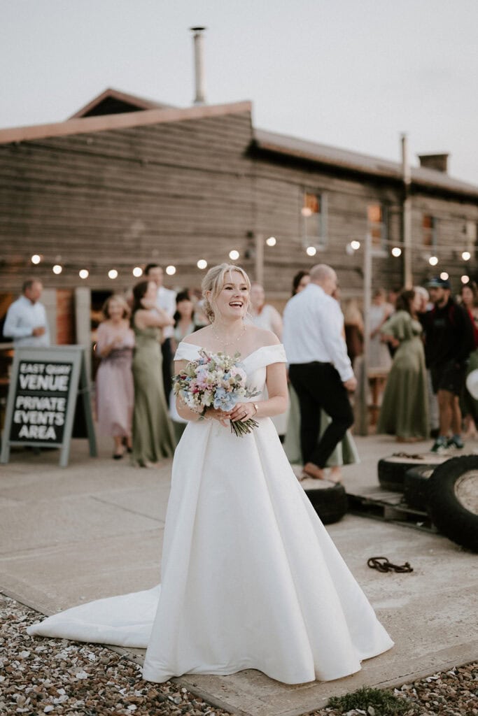 A bride in a white off-the-shoulder gown holds a bouquet of pastel flowers, smiling. Behind her, guests in formal attire gather outside the East Quay Venue, a rustic building adorned with string lights. A sign in the background reads "Venue Private Events Area," setting the perfect wedding scene. Image by Pearce Wedding Photography.