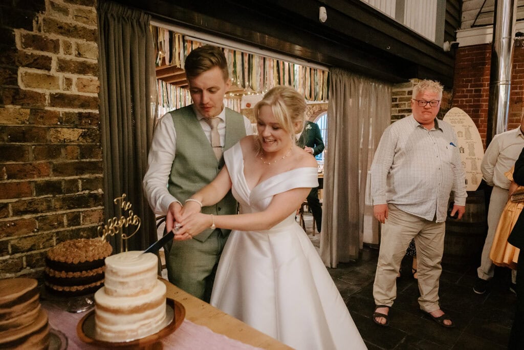 A couple in wedding attire smiles while cutting a tiered cake together at the East Quay Venue. They are indoors, with colorful curtains in the background. An older man stands to the right, watching the scene unfold. Several other cakes are visible on the table, adding to the festive atmosphere. Image by Pearce Wedding Photography.