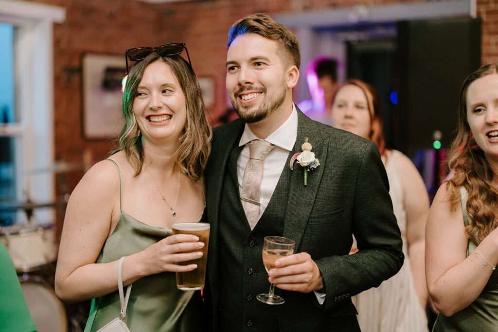 A smiling couple poses together at the lively wedding. The woman in a green dress holds a drink, while the man in a suit with a boutonniere holds a glass. Other attendees are visible in the warmly lit background of the East Quay Venue. Image by Pearce Wedding Photography.