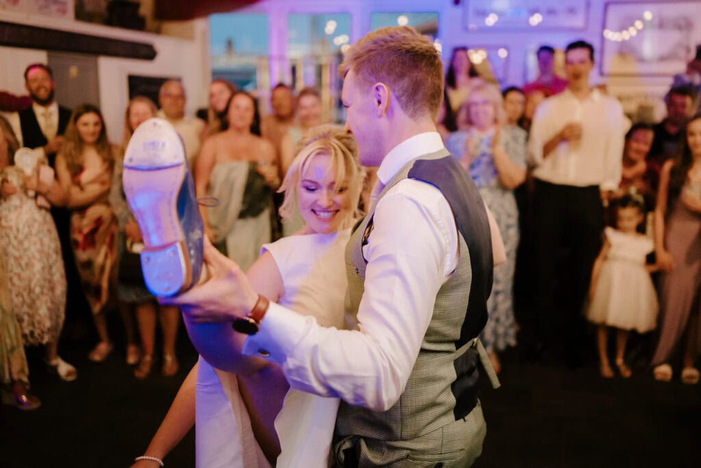 At the East Quay Venue, a couple dances joyfully at their wedding reception. The groom lifts the bride, who smiles as she playfully holds her shoe. Guests watch and smile in the warmly lit, festive atmosphere. Image by Pearce Wedding Photography.