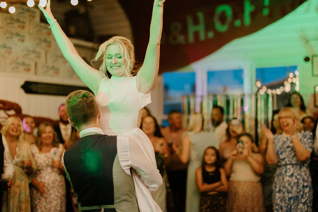 A bride is joyfully lifted into the air by her partner on the dance floor at East Quay Venue, surrounded by cheering guests. The room, adorned with twinkling lights and a festive banner, exudes a lively wedding celebration atmosphere. Image by Pearce Wedding Photography.