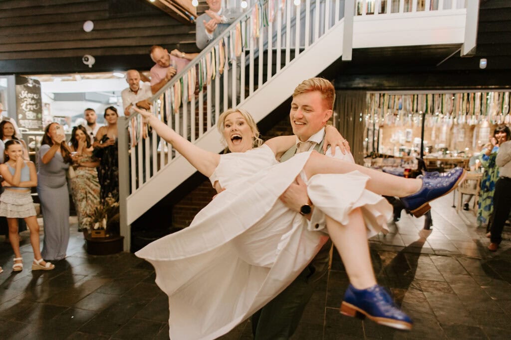 A joyful couple is dancing indoors at East Quay Venue, with the man lifting the woman in her white dress and blue shoes. He is in a suit. Guests, smiling near the staircase, watch as colorful streamers hang overhead, celebrating their wedding day. Image by Pearce Wedding Photography.