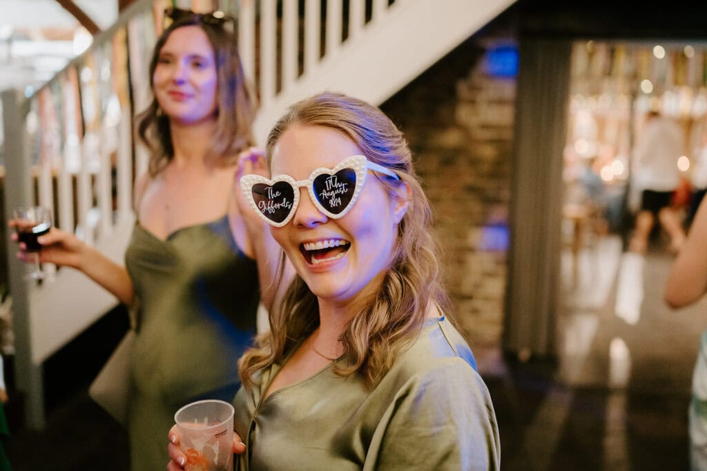 A person in a green dress smiles widely at a vibrant wedding party, wearing heart-shaped sunglasses with playful text. Another guest holds a drink and smiles. The celebration unfolds indoors at the East Quay Venue, with its iconic staircase and festive lighting creating an enchanting ambiance. Image by Pearce Wedding Photography.