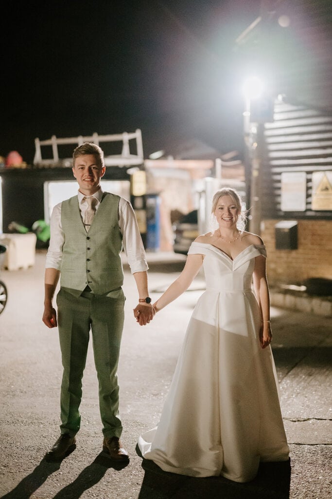 A couple stands hand in hand outdoors at night, possibly celebrating their wedding. The man dons a light green vest with a white shirt, while the woman wears an off-the-shoulder white gown. Both smile warmly, illuminated by a bright light behind them, reminiscent of the enchanting East Quay Venue ambiance. Image by Pearce Wedding Photography.