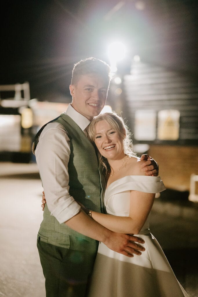 A smiling couple in wedding attire embrace outdoors at night at the East Quay Venue, illuminated by a bright light in the background. The bride wears a white dress, and the groom sports a vest and shirt. They appear happy and content. Image by Pearce Wedding Photography.