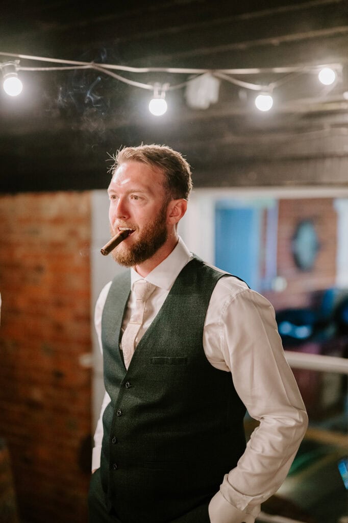 A man in a formal vest and tie stands under string lights at the East Quay Venue, holding a lit cigar. The dimly lit room, perfect for a wedding, showcases rustic brick and wooden walls. Image by Pearce Wedding Photography.