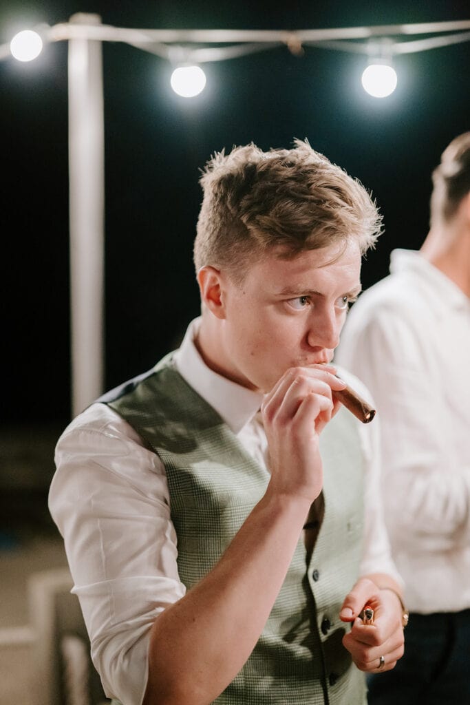 Under the twinkling string lights at the East Quay Venue, a person in a crisp white shirt and vest savors a cigar with a thoughtful expression, reminiscent of wedding nights full of quiet moments. Another figure is partially visible in the background, adding to the evening's charm. Image by Pearce Wedding Photography.