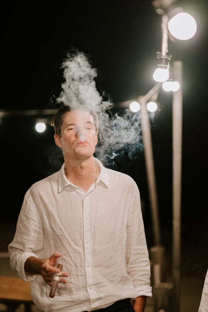 A person in a white shirt is exhaling smoke while holding a cigarette at the East Quay Venue. They are standing outdoors at night, surrounded by glowing string lights, perhaps taking a quiet moment during the wedding festivities. Image by Pearce Wedding Photography.