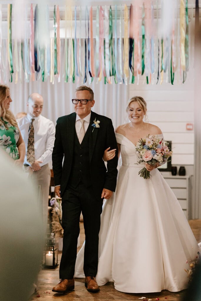 A bride in a white dress beams as she walks arm in arm with a man in a suit down the decorated aisle of the East Quay Venue. She holds a colorful bouquet, while guests and vibrant ribbons hang above, celebrating this beautiful wedding. Image by Pearce Wedding Photography.