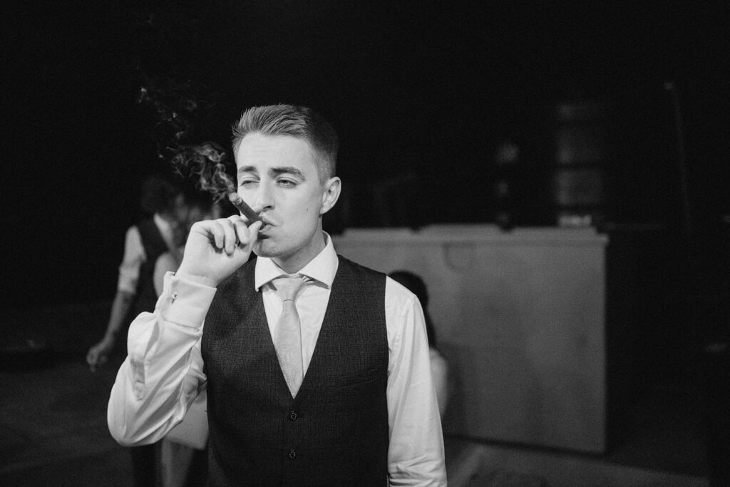 A man in formal attire, wearing a vest and tie, is smoking a cigar at a wedding. The black and white image captures the relaxed atmosphere at the East Quay Venue, with another person blurred in the background. Image by Pearce Wedding Photography.