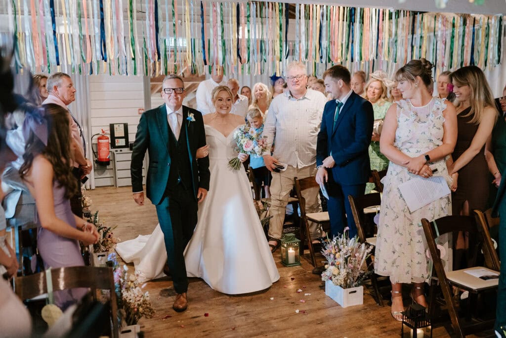 A bride in a white gown walks down the aisle with an older man, likely her father. Guests at the East Quay venue line the wooden walkway, smiling and watching. Colorful ribbons hang from the ceiling, adding a festive touch to this beautiful wedding ceremony. Image by Pearce Wedding Photography.