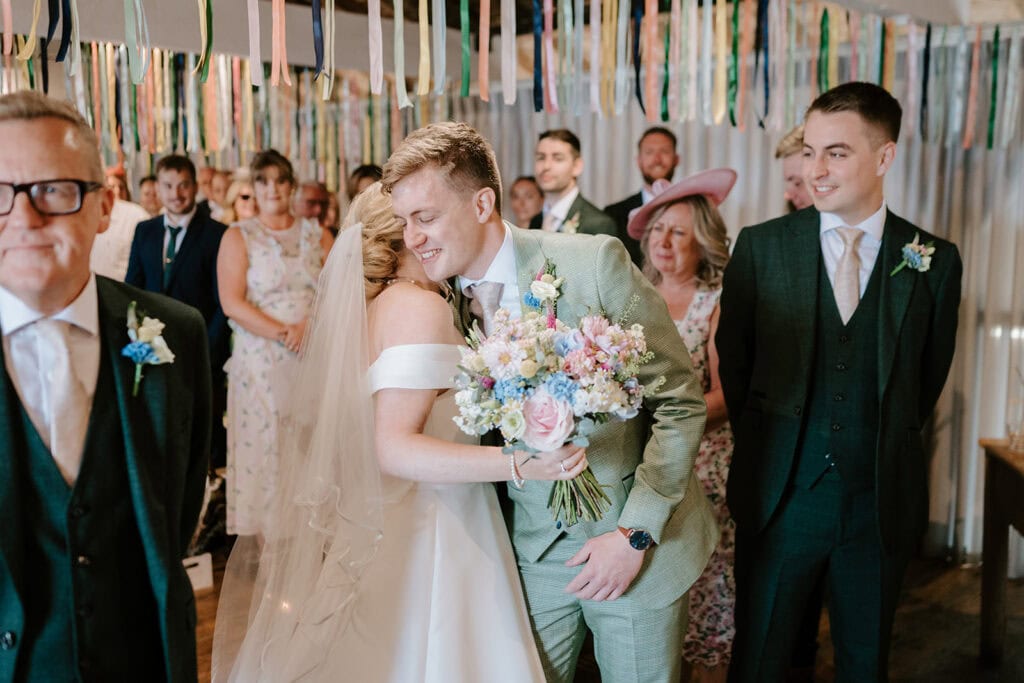 At the East Quay Venue, a bride and groom share a joyful hug in a beautifully decorated room with colorful streamers. The groom is in a light green suit, while the bride holds a bouquet of pastel flowers. Wedding guests look on with smiles. Image by Pearce Wedding Photography.