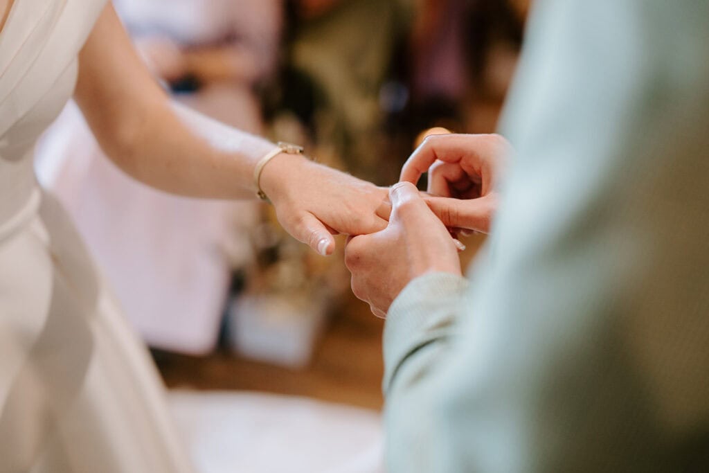 A person in a wedding dress at East Quay Venue is having a ring placed on their finger by someone in a green sweater, symbolizing the joyous union. The focus remains on the hands, set against a beautifully blurred background. Image by Pearce Wedding Photography.