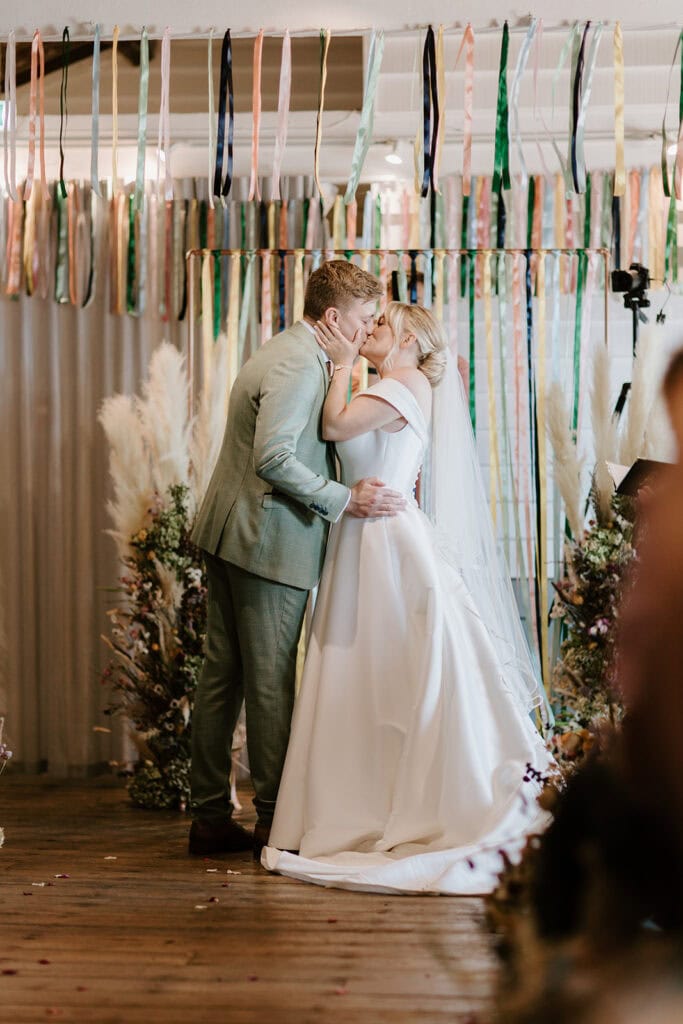 At the East Quay Venue, a couple shares a tender kiss during their wedding ceremony. The bride, stunning in her white gown and long veil, stands beside the groom in his light gray suit, surrounded by decorative ribbons and exquisite floral arrangements. Image by Pearce Wedding Photography.
