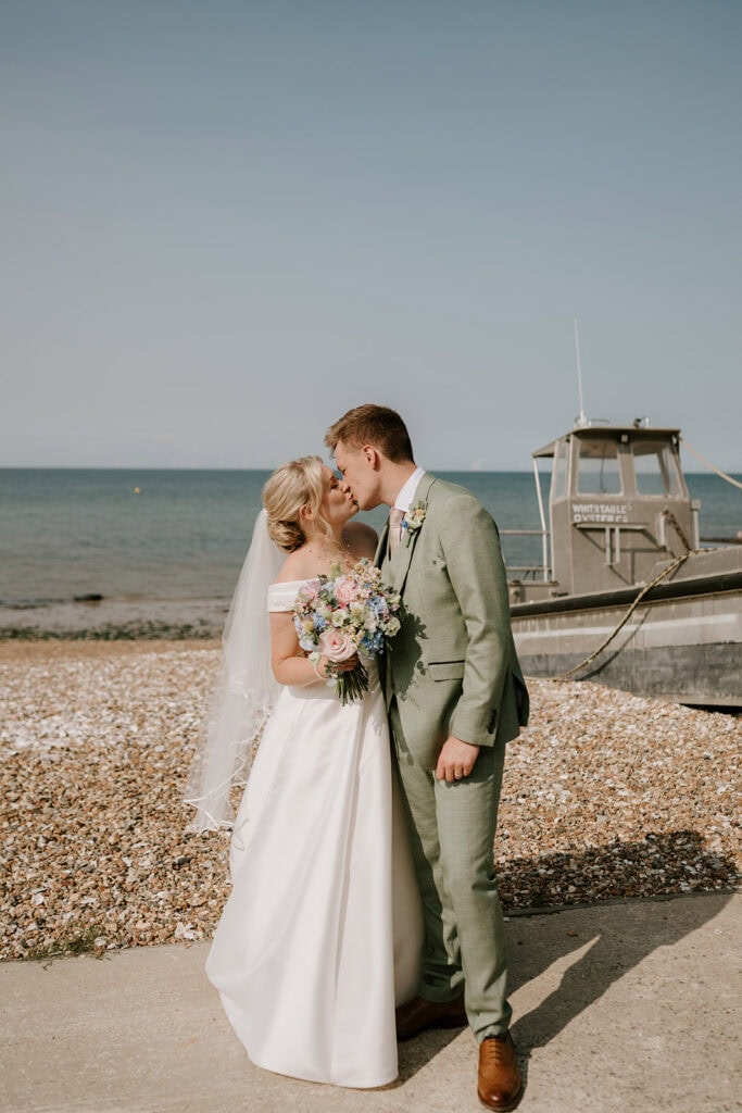 On a pebbled beach, a bride and groom share a tender kiss. The bride clutches a bouquet of flowers while the groom stands proudly in his green suit. A boat drifts softly in the background against the clear blue sky, capturing the essence of their wedding at the East Quay venue. Image by Pearce Wedding Photography.