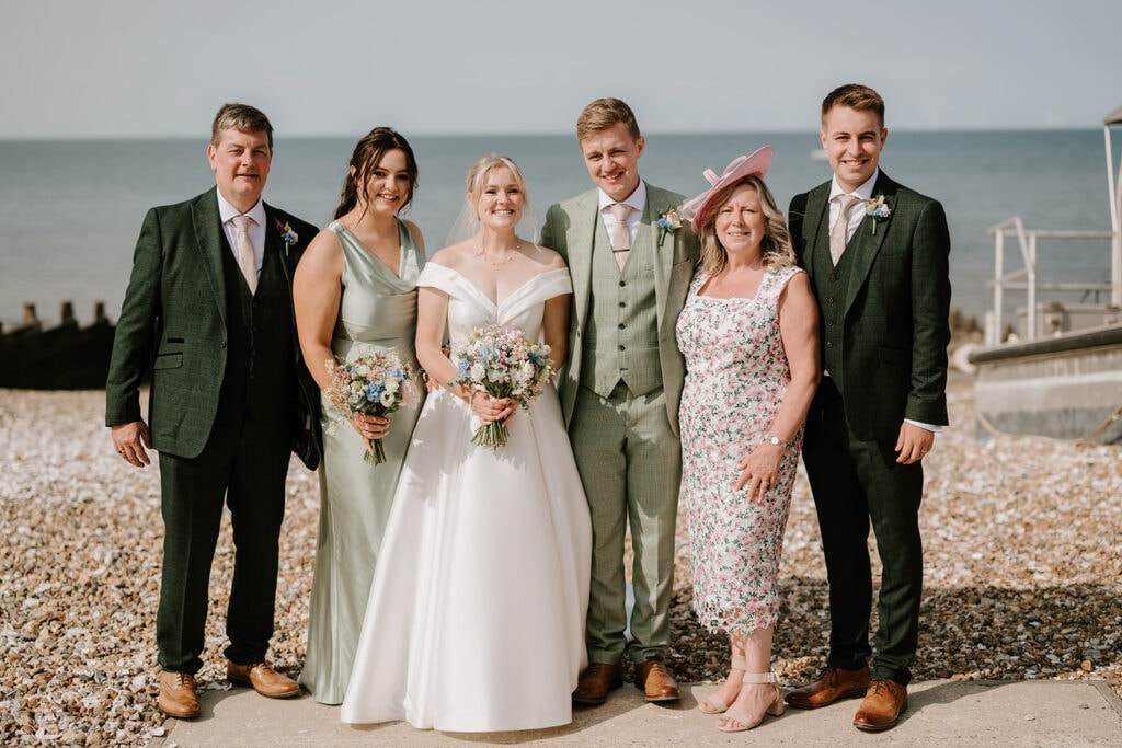 A wedding party gathers on a pebble beach near the East Quay Venue. The bride and groom stand at the center, surrounded by three other adults. The women wear dresses, while the men don suits, with a boat and the sea providing a picturesque backdrop. Image by Pearce Wedding Photography.