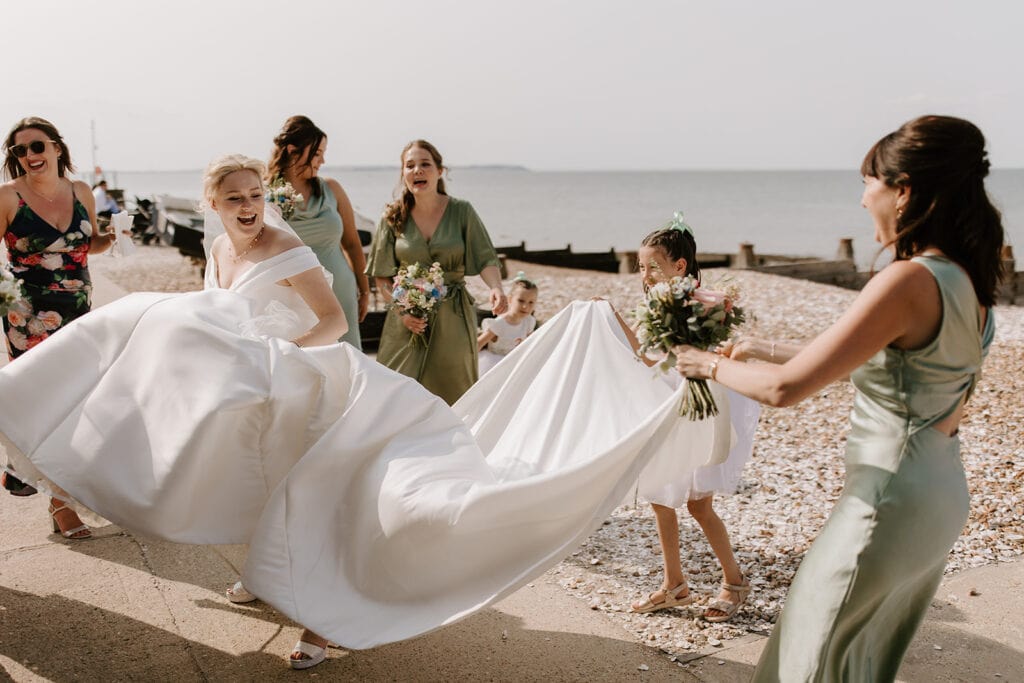A joyful bride in a flowing white dress is surrounded by bridesmaids in green dresses and a flower girl at the stunning East Quay Venue. They stand on a pebbled beach with the sea as their backdrop, under a clear sky. Everyone is smiling and laughing, holding bouquets, capturing the wedding's delight. Image by Pearce Wedding Photography.