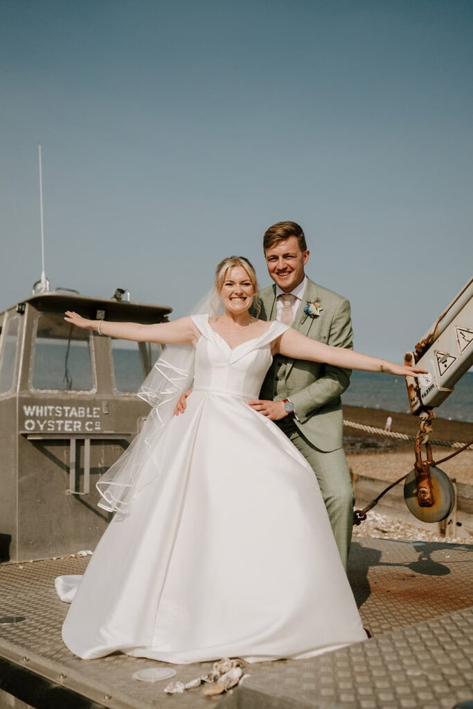 A bride in a white dress and a groom in a light green suit stand joyfully at the East Quay Venue by the sea. The bride spreads her arms wide, smiling, while the groom stands behind her. A boat labeled "Whitstable Oyster Co" is in the background of their enchanting wedding scene. Image by Pearce Wedding Photography.