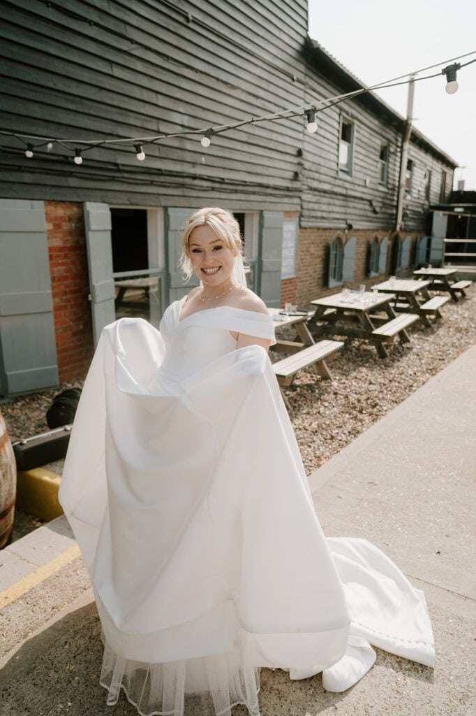 A smiling bride in an off-shoulder white wedding dress holds up her gown outdoors at the East Quay Venue. She stands by wooden benches and a rustic building with blue shutters, under string lights. Her hair is styled in an elegant updo, perfect for the wedding setting. Image by Pearce Wedding Photography.