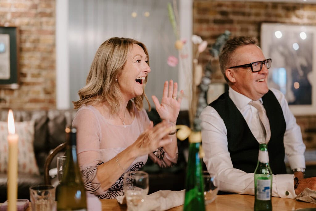 A smiling woman and man sit at a restaurant table at an East Quay Venue, engaging in conversation. The woman is wearing a light-colored top and the man is in a suit. There are food and drinks on the table, with framed pictures adorning the brick wall background, adding charm to their delightful gathering. Image by Pearce Wedding Photography.