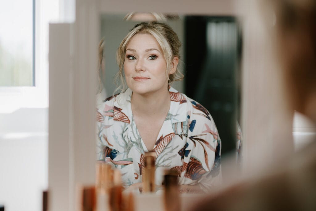 A person with blonde hair is reflected in a mirror, wearing a top with a colorful floral pattern. In preparation for their wedding at the East Quay Venue, they are seen applying makeup, with several cosmetic items visible in the foreground on a table. Image by Pearce Wedding Photography.