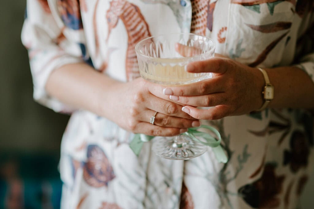 A person in a patterned shirt gracefully holds a large, elegant glass with both hands, perhaps at a chic wedding reception at the East Quay Venue. A ring and watch glint subtly on their left hand and wrist, adding to the sophisticated atmosphere. Image by Pearce Wedding Photography.